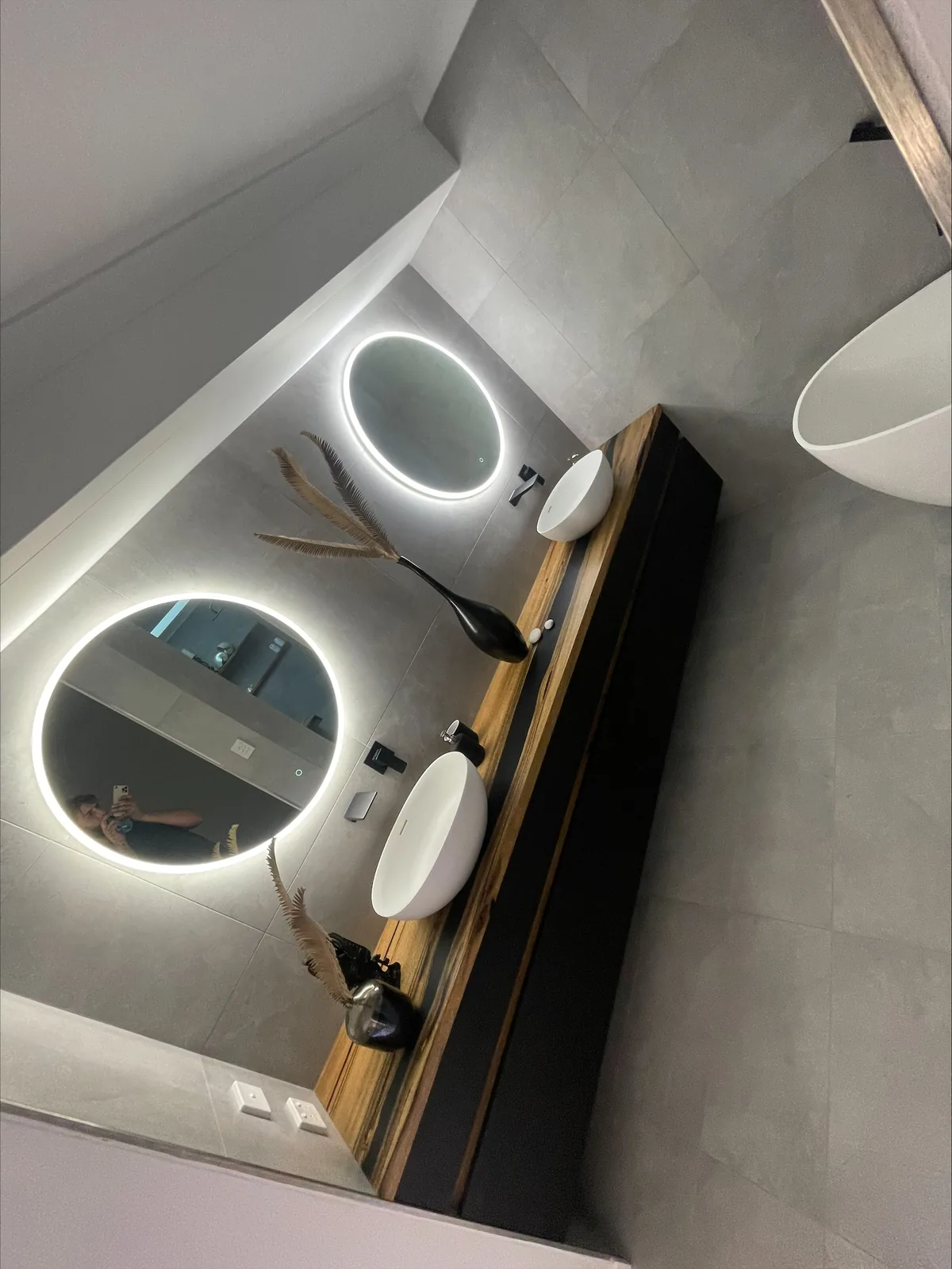 Floating black vanity with twin basins and round backlit LED mirrors against a concrete-look tiled wall